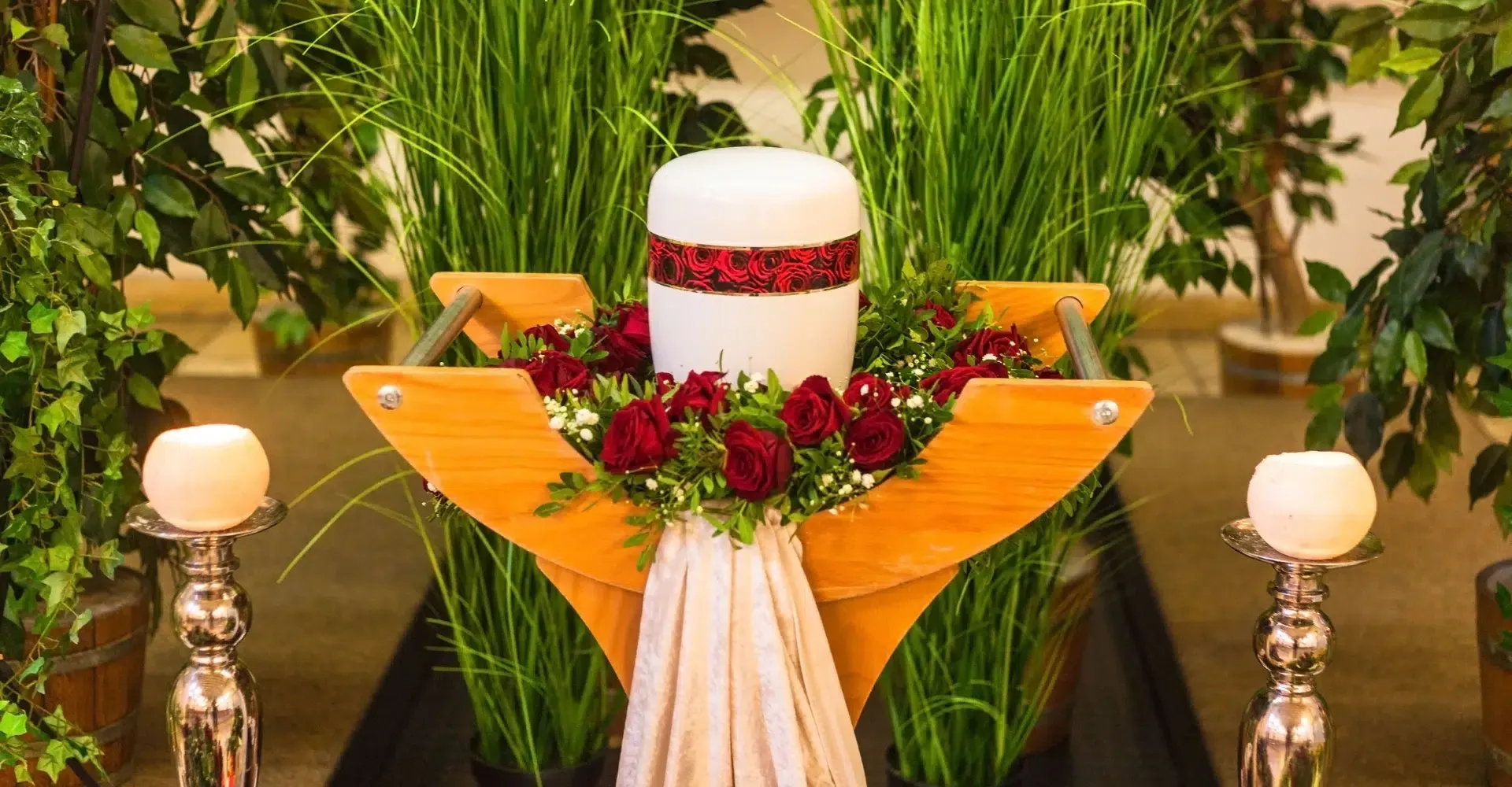 A white urn surrounded by red roses and candles on a wooden stand.