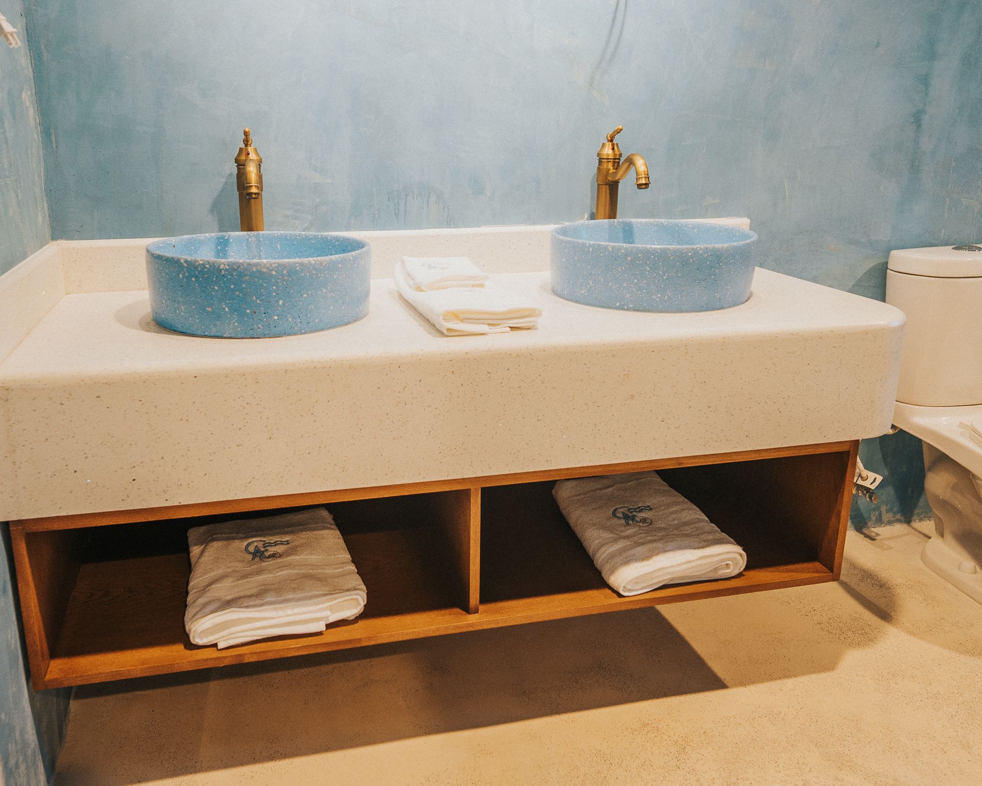 Baño con dos lavabos azules, grifos dorados y un mueble de madera que sostiene toallas contra una pared azul.