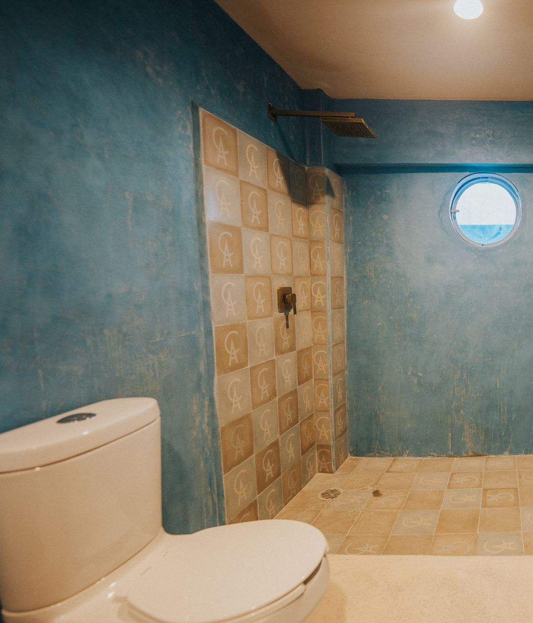 Baño con paredes azules, ducha de azulejos e inodoro blanco. Incluye una ventana circular.