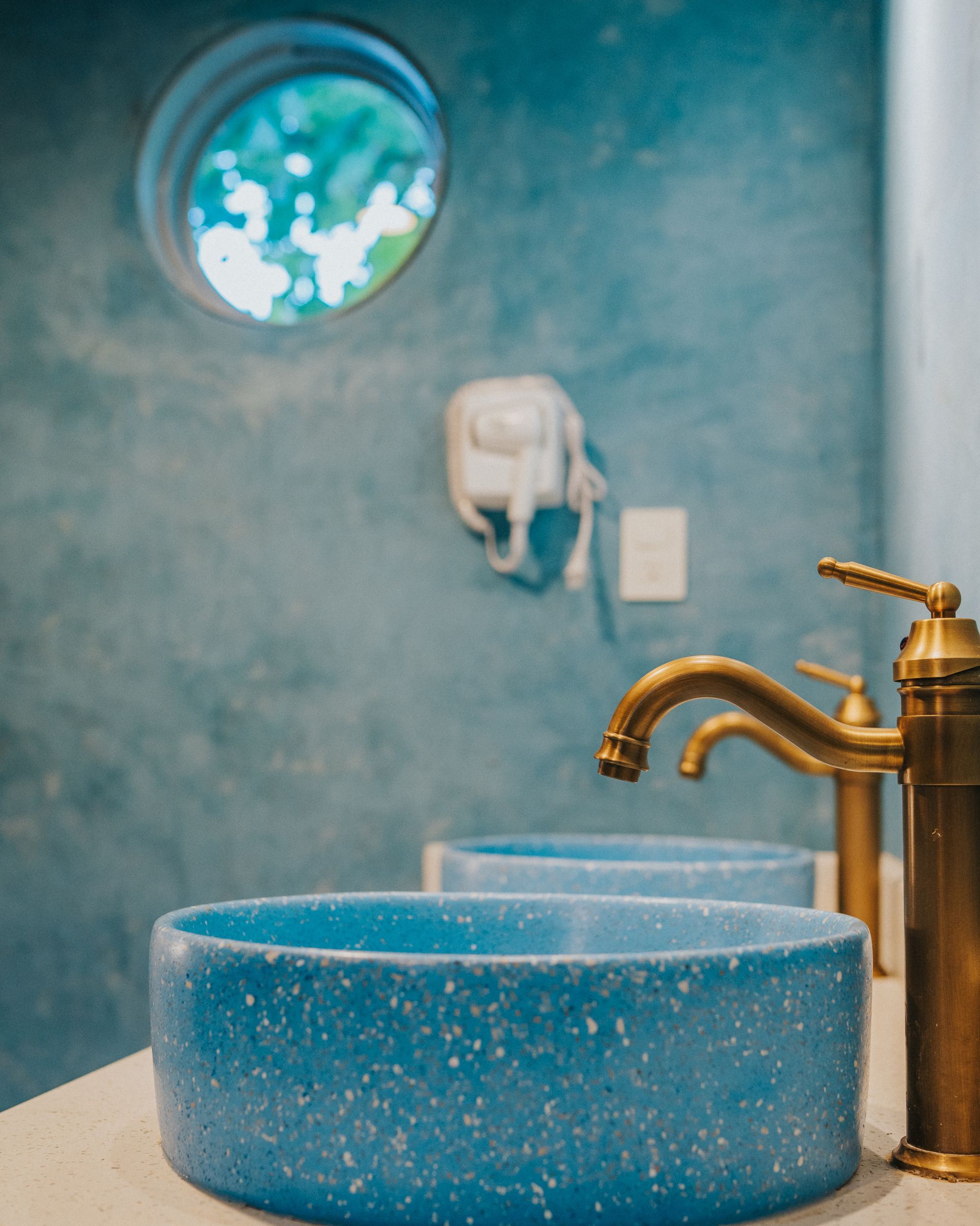 Baño azul con lavabos redondos, grifos de latón y ventana de ojo de buey.