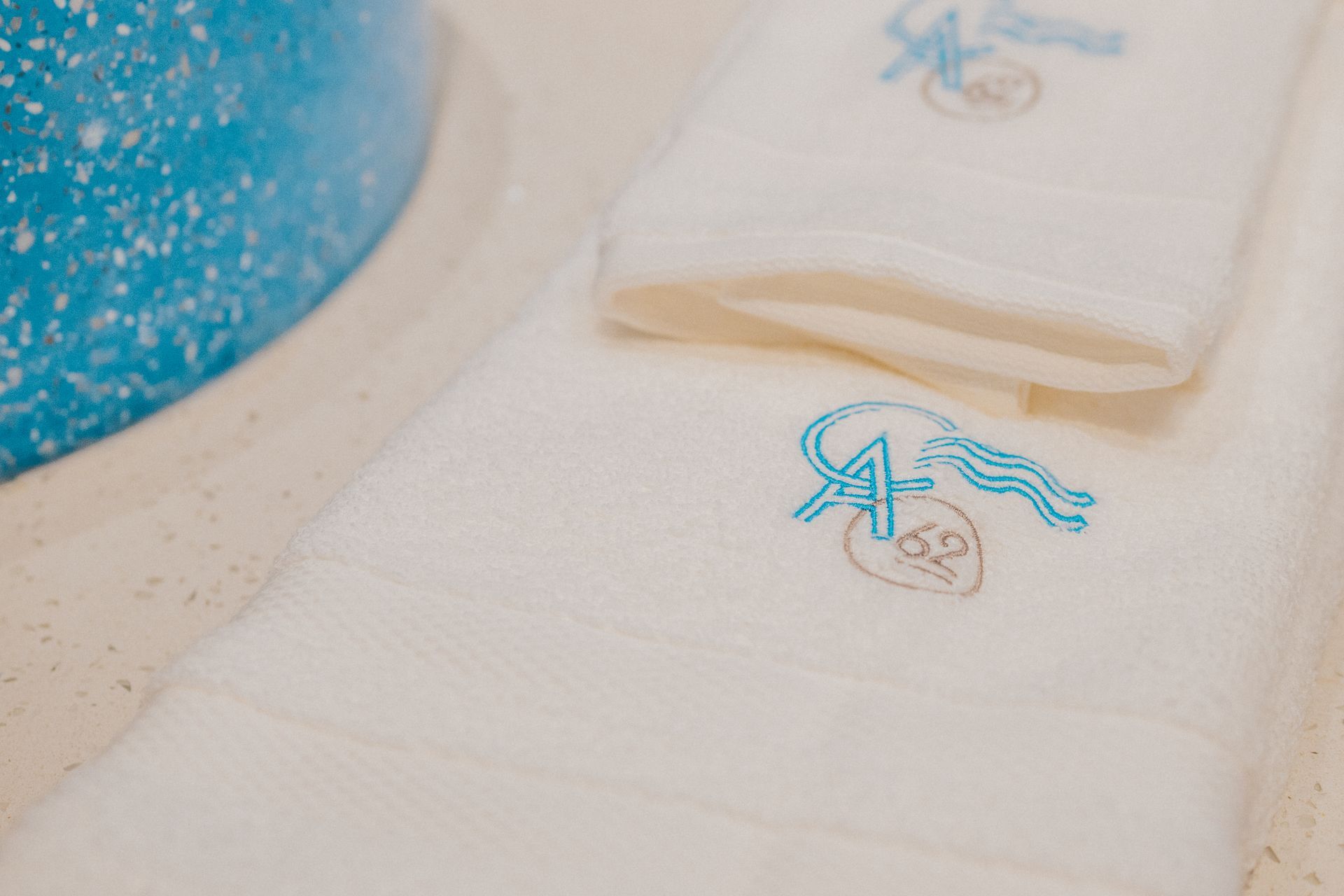 Toallas blancas con logo azul bordado, colocadas junto a un lavabo azul moteado.