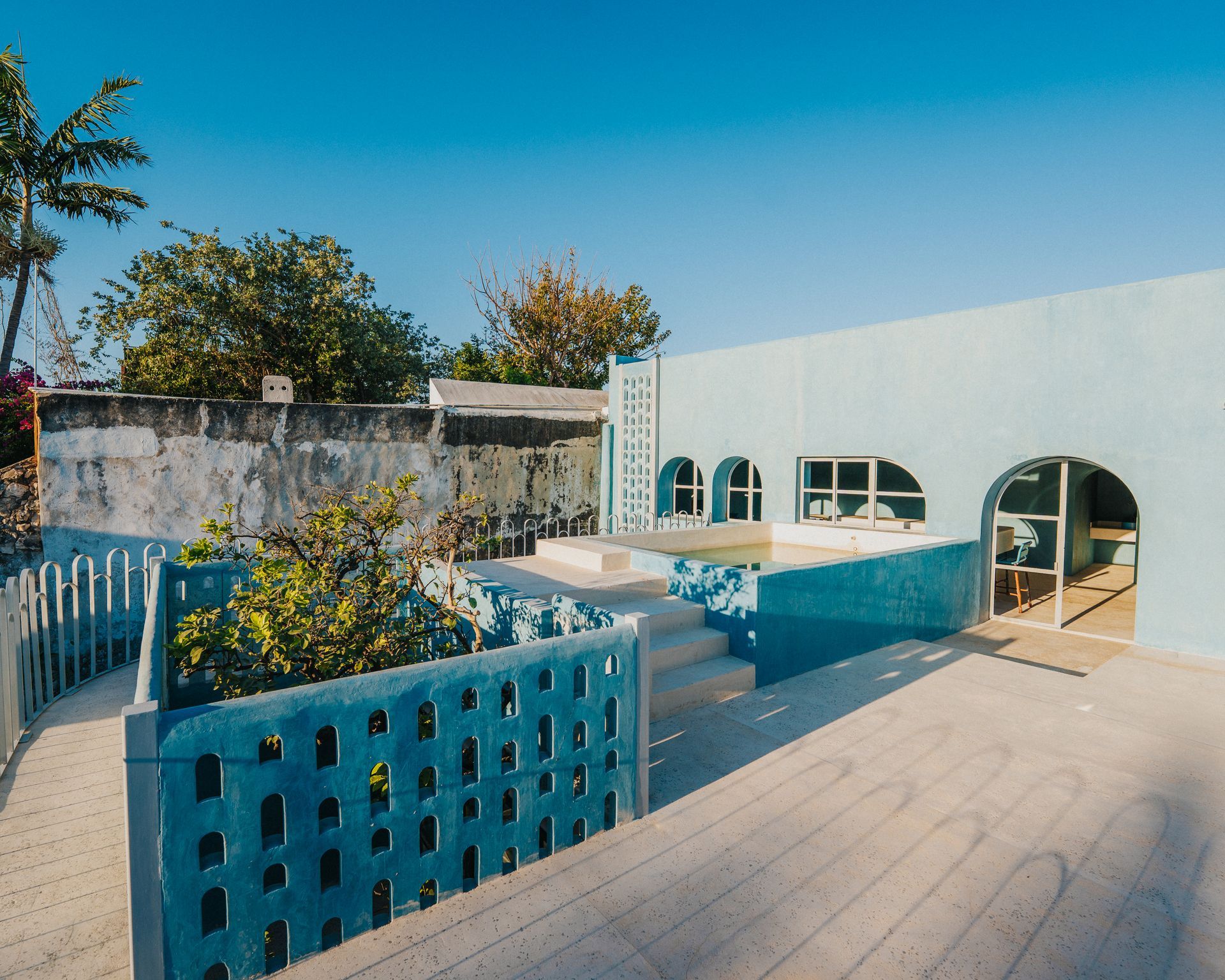 Edificio pintado de azul con ventanas arqueadas, piscina y patio bajo un cielo azul claro.
