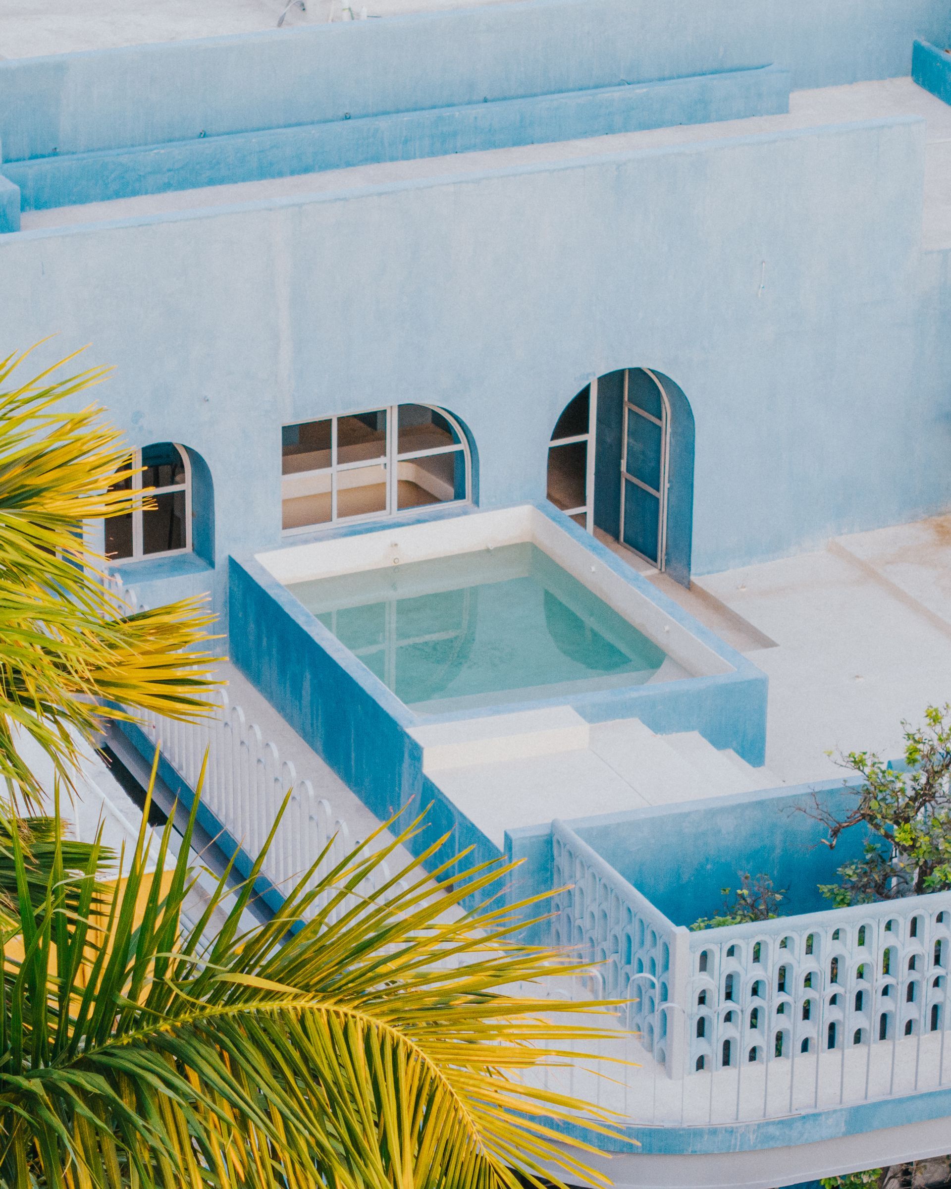 Piscina azul en la azotea, edificio con ventanas arqueadas. Hojas de palmera enmarcan la escena.