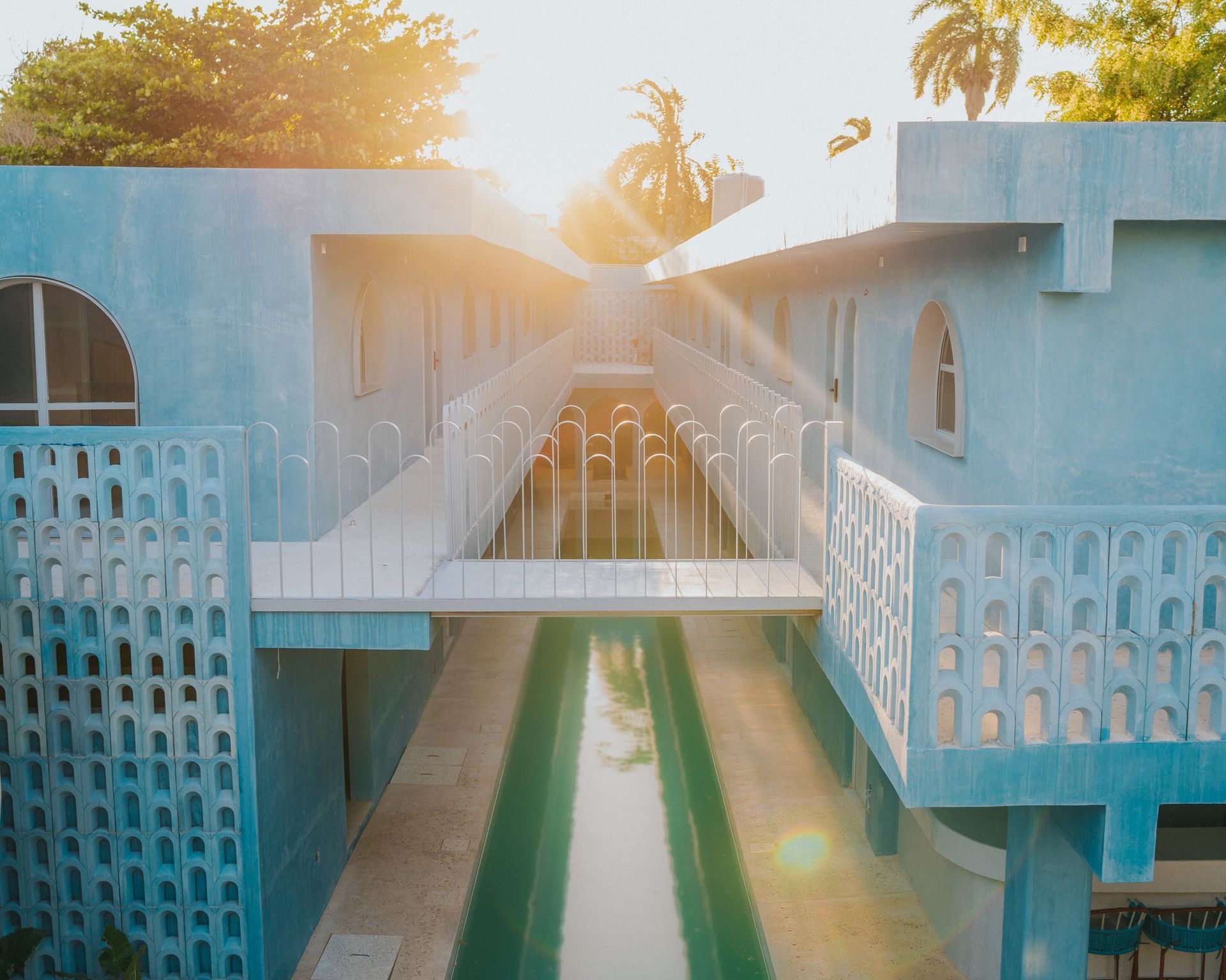 Edificio modernista azul con una larga piscina, un puente de conexión y una luz solar brillante.