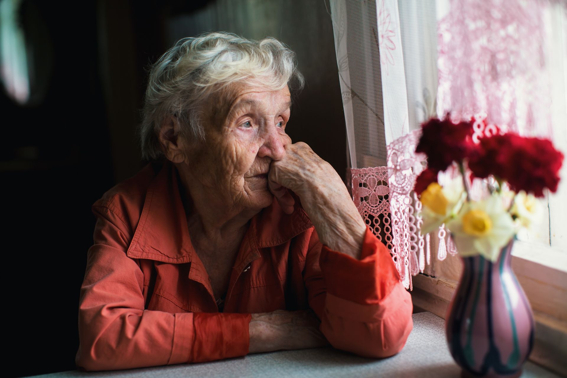 An elderly woman is sitting at a table looking out of a window.