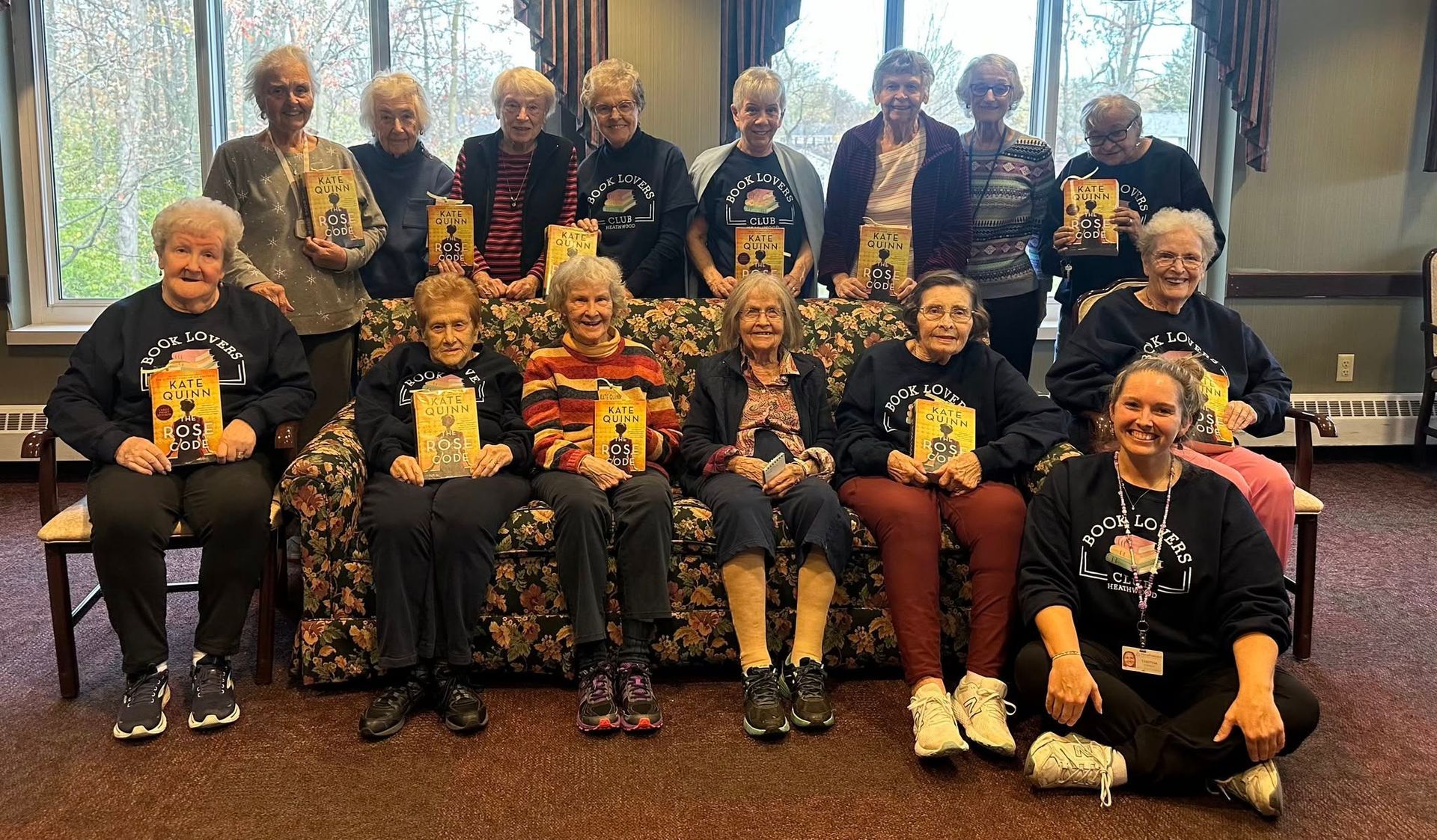Group of seniors holding books, posing indoors. They are seated in front of a window, wearing matching shirts.