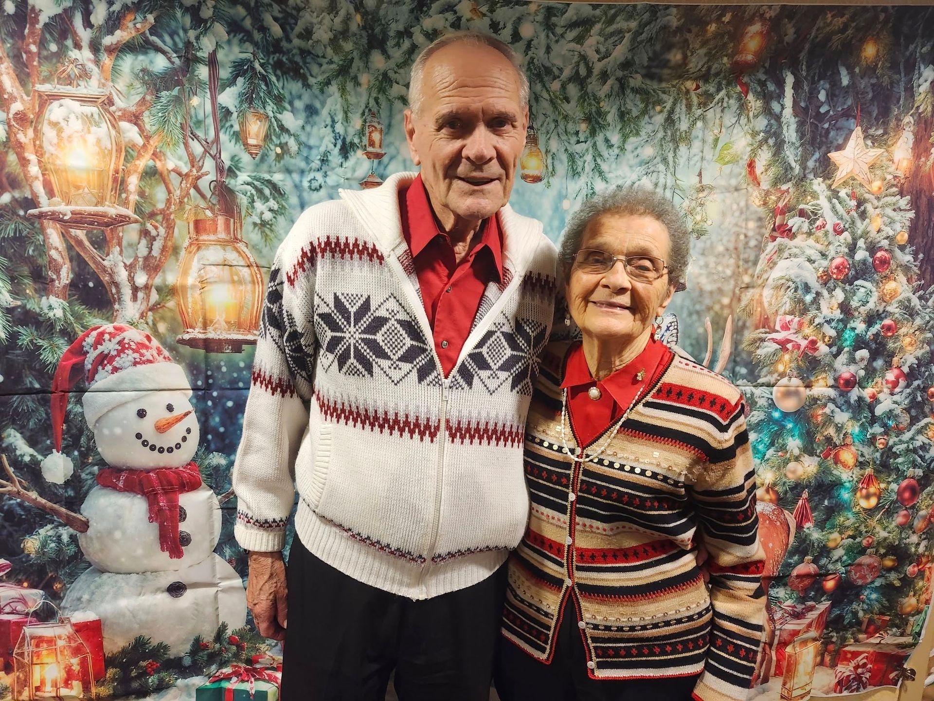 Woman and man in a wheelchair smiling outdoors, holding hands. Man wearing sunglasses and a blue shirt. Green foliage and flowers in the background.