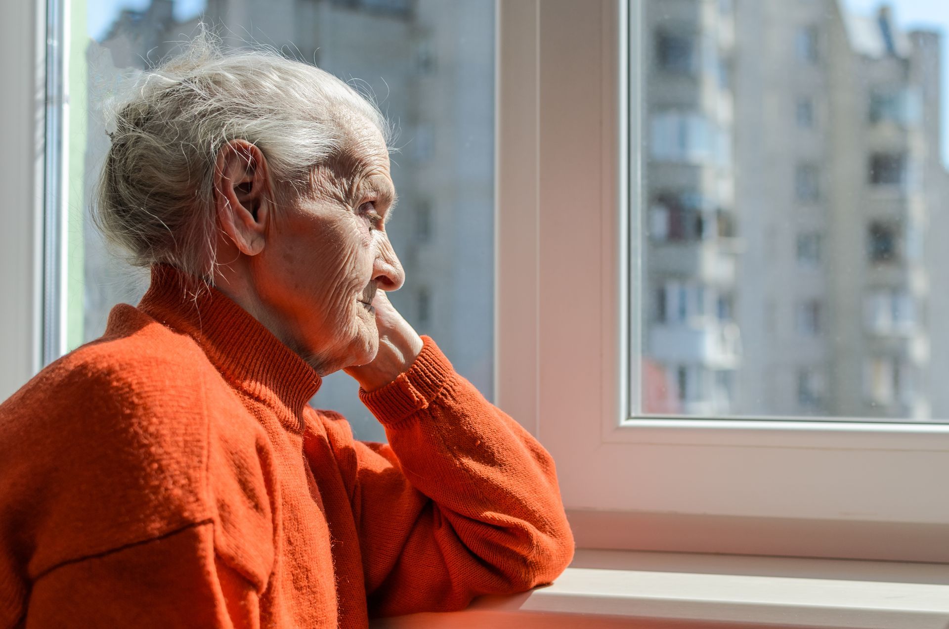 An elderly woman in an orange sweater is looking out of a window.