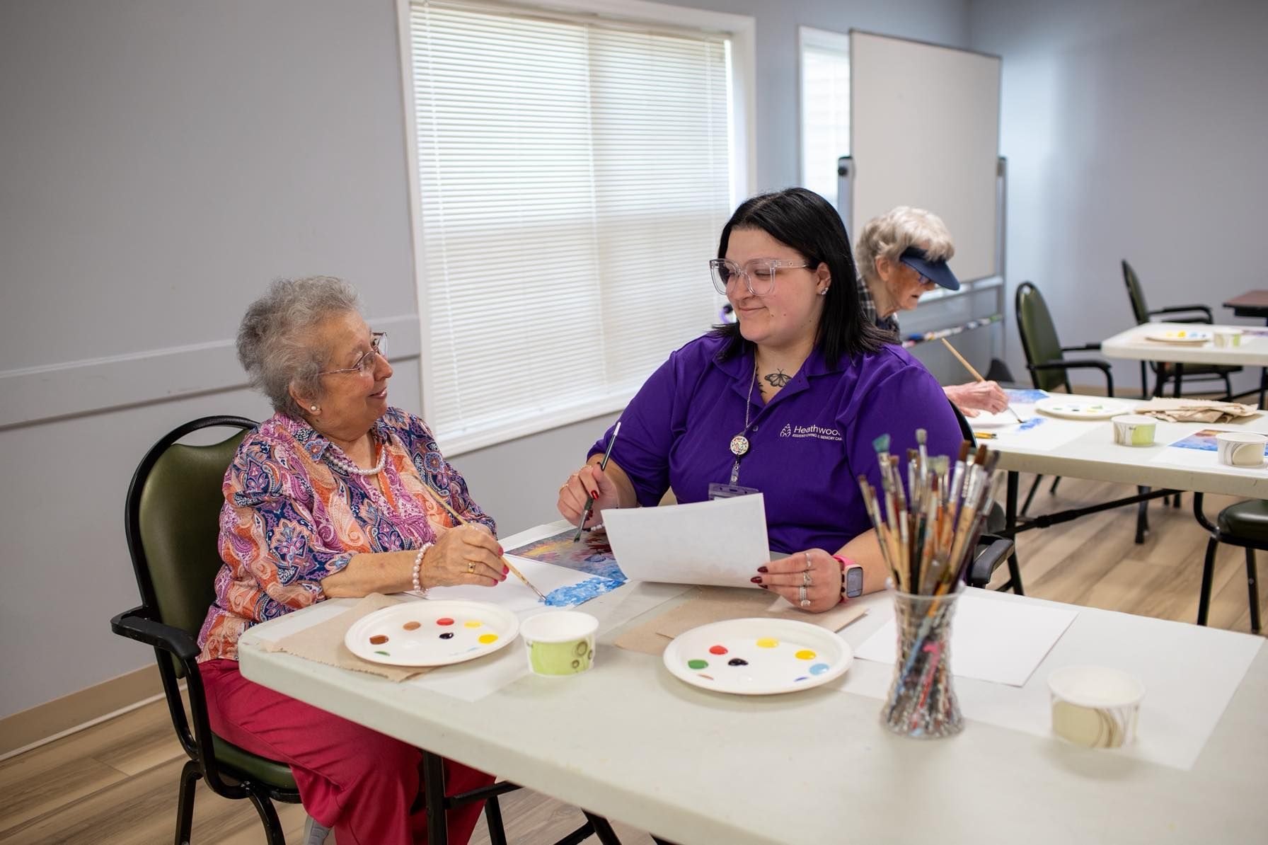 Woman in purple shirt assists elderly woman at an art class table.