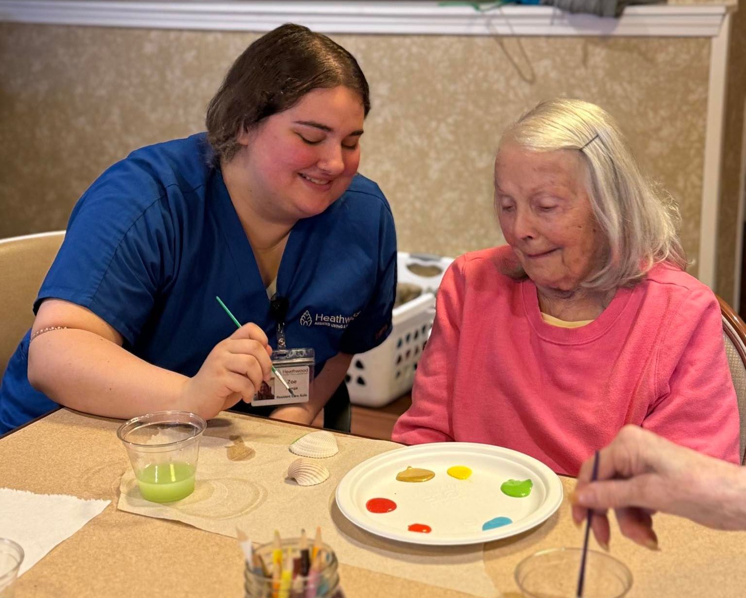 A caregiver and elderly woman paint at a table. The caregiver smiles, and both look at the project.