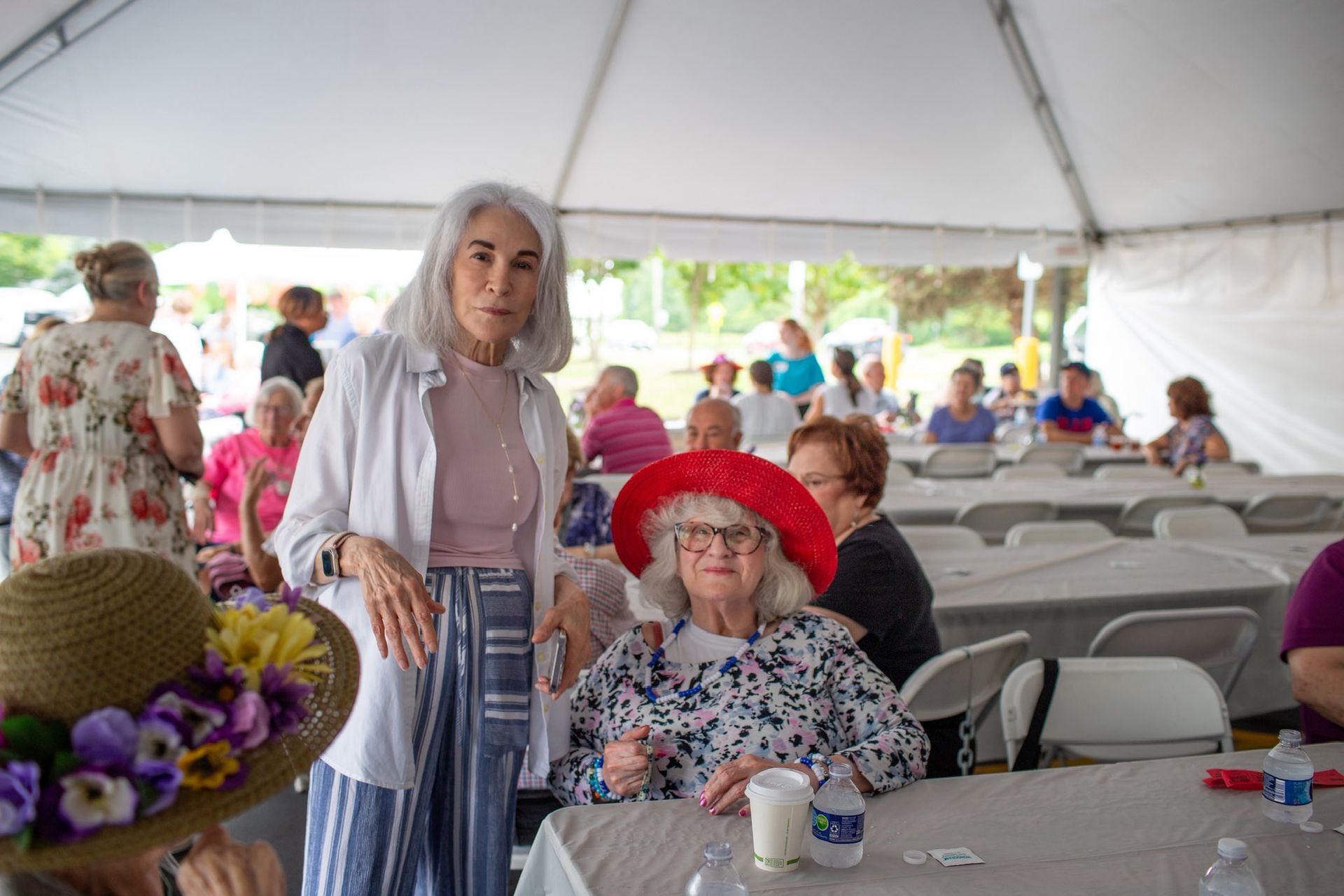 Two women under a tent, one with a red hat, others seated at tables.
