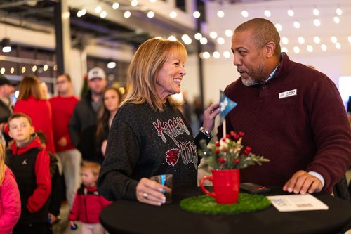 Two people talk and smile at a small high-top table in a festive, softly lit room with string lights.