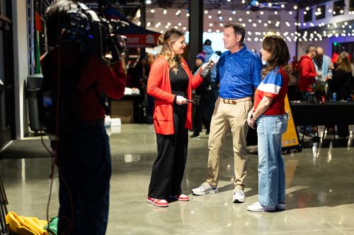 A reporter holds a microphone interviewing a man and woman in a brightly lit, industrial-style indoor event space.