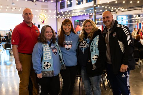 Five people smiling for a photo indoors, wearing soccer fan apparel and scarves in a decorated event space.
