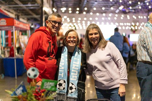 Three people pose smiling at an indoor event, with the person in the middle wearing a blue and white soccer-themed scarf.