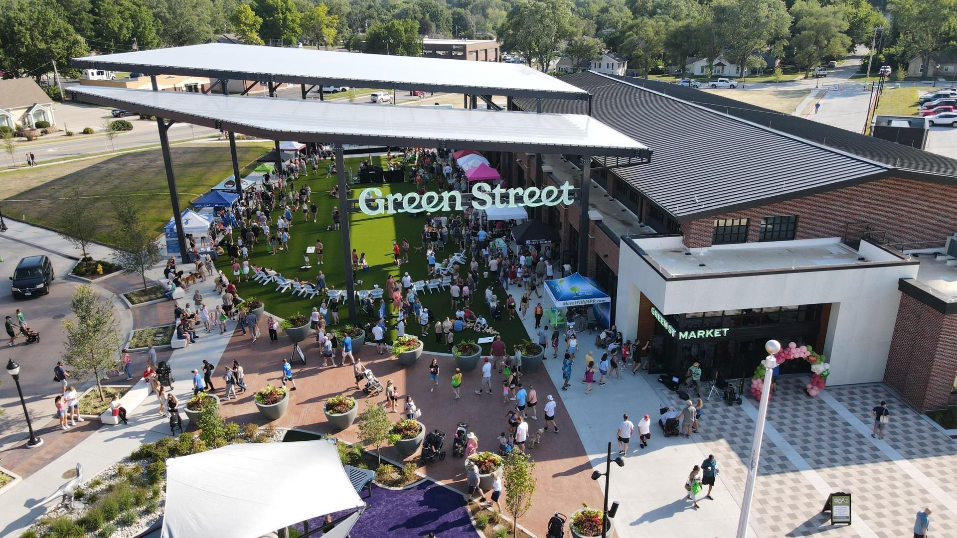 Green Street market, outdoor event with many people. Green sign. Building with angled roof, sunny day.