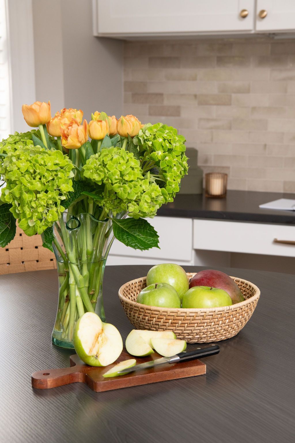 A vase of flowers sits on a kitchen counter next to a basket of apples.