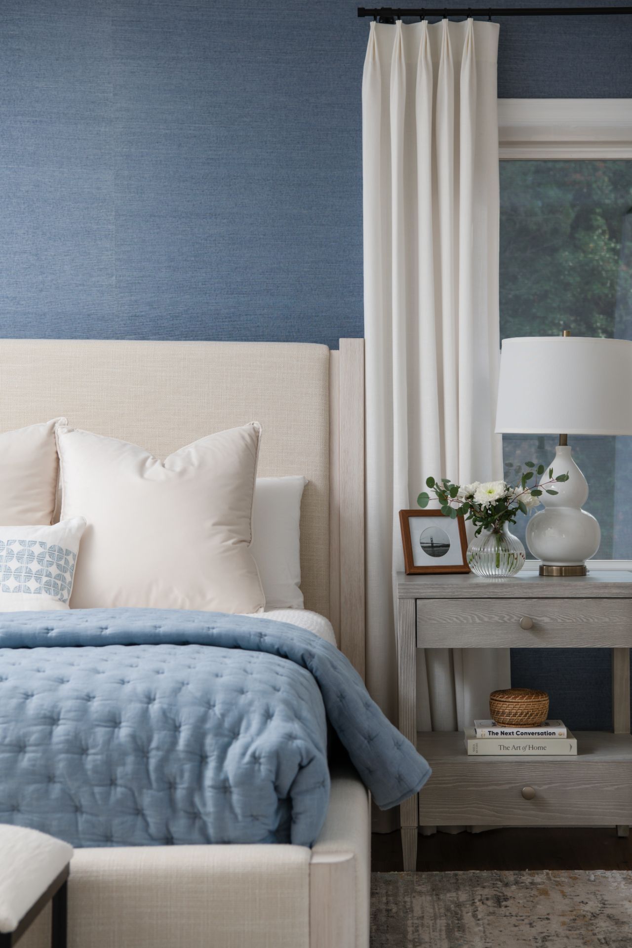 Bedroom with blue walls, a cream-colored bed, and a wooden nightstand. A window with white curtains is visible | Charlotte, NC | An Inspiring Home