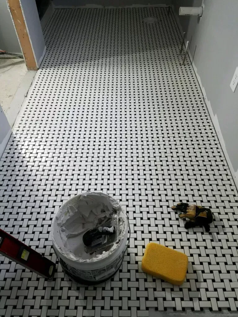 A bucket of grout is sitting on a tiled floor next to a yellow sponge.