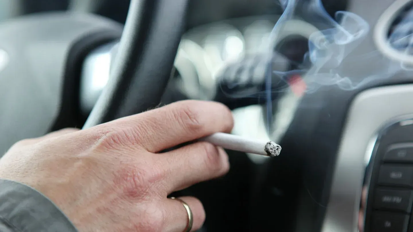 A hand wearing a gold ring holds a lit cigarette with rising smoke inside a car.