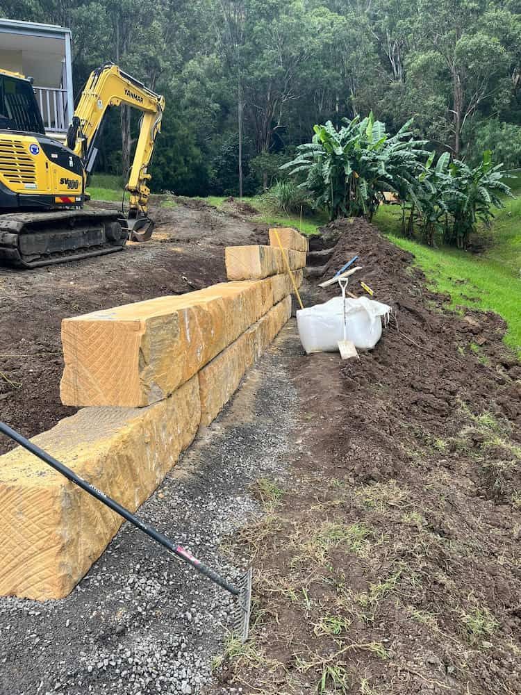 A Yellow Excavator is Working on a Stone Wall — BeCivil in Huskisson, NSW