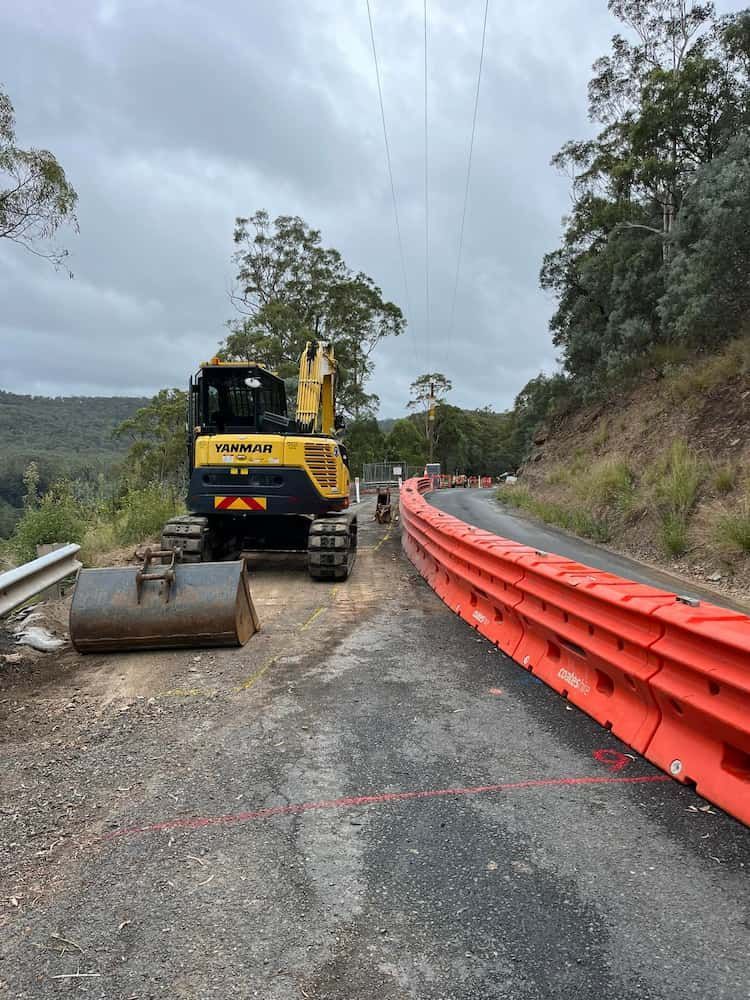 A Yellow and Blue Excavator is Sitting on the Side of a Road Next to an Orange Barrier — BeCivil in Ulladulla, NSW