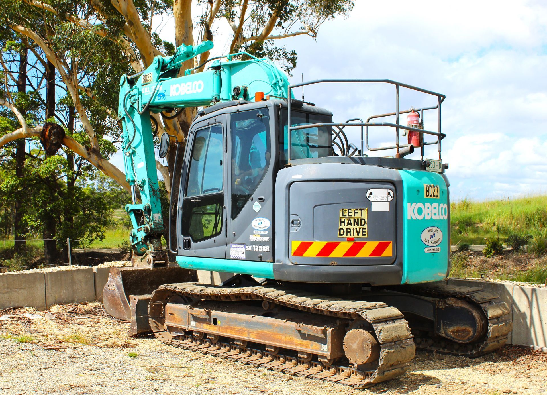 A blue and gray excavator is parked in a gravel lot next to a tree — Blue Dog Landscape Supplies In Newee Creek, NSW