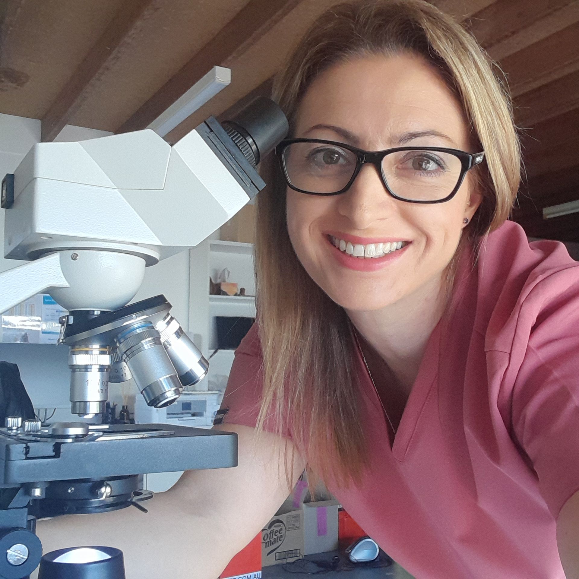A woman wearing glasses is smiling in front of a microscope
