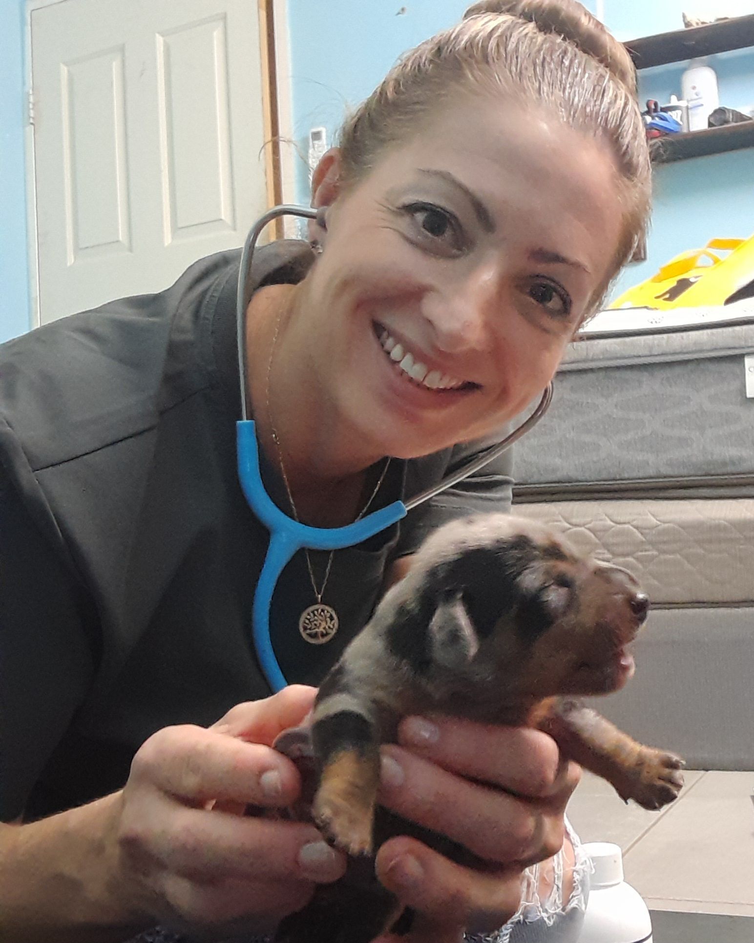 A woman is holding a puppy and smiling while wearing a stethoscope.