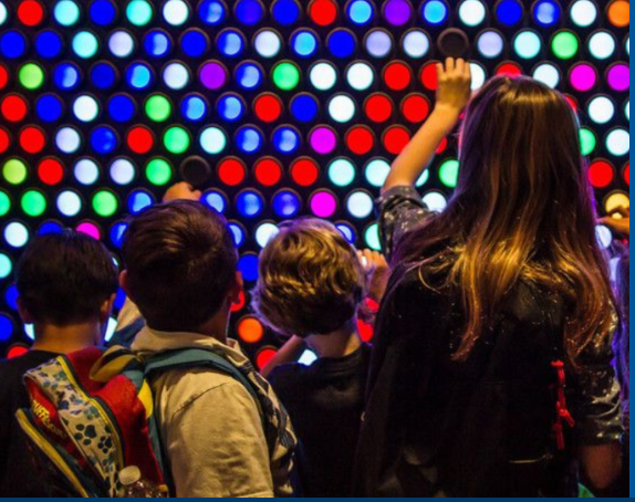 A group of children are standing in front of a wall of colorful lights