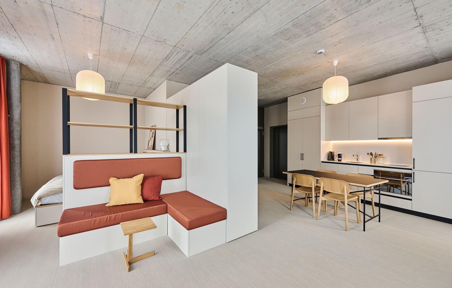 Modern studio apartment with a red sofa bed, dining table, and kitchen under a concrete ceiling