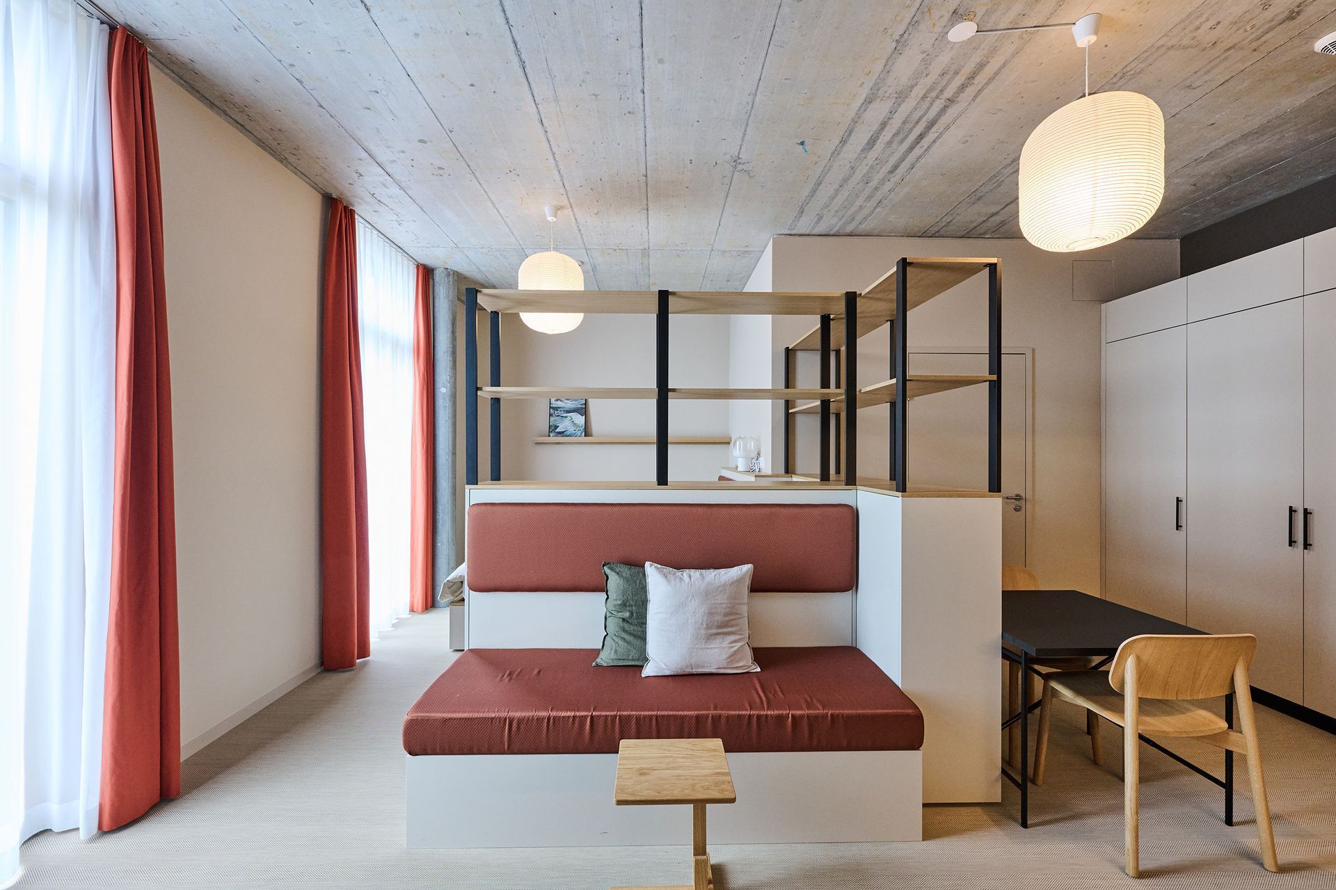 Modern studio apartment with red sofa, desk, and built-in wardrobes under a lofted shelf.
