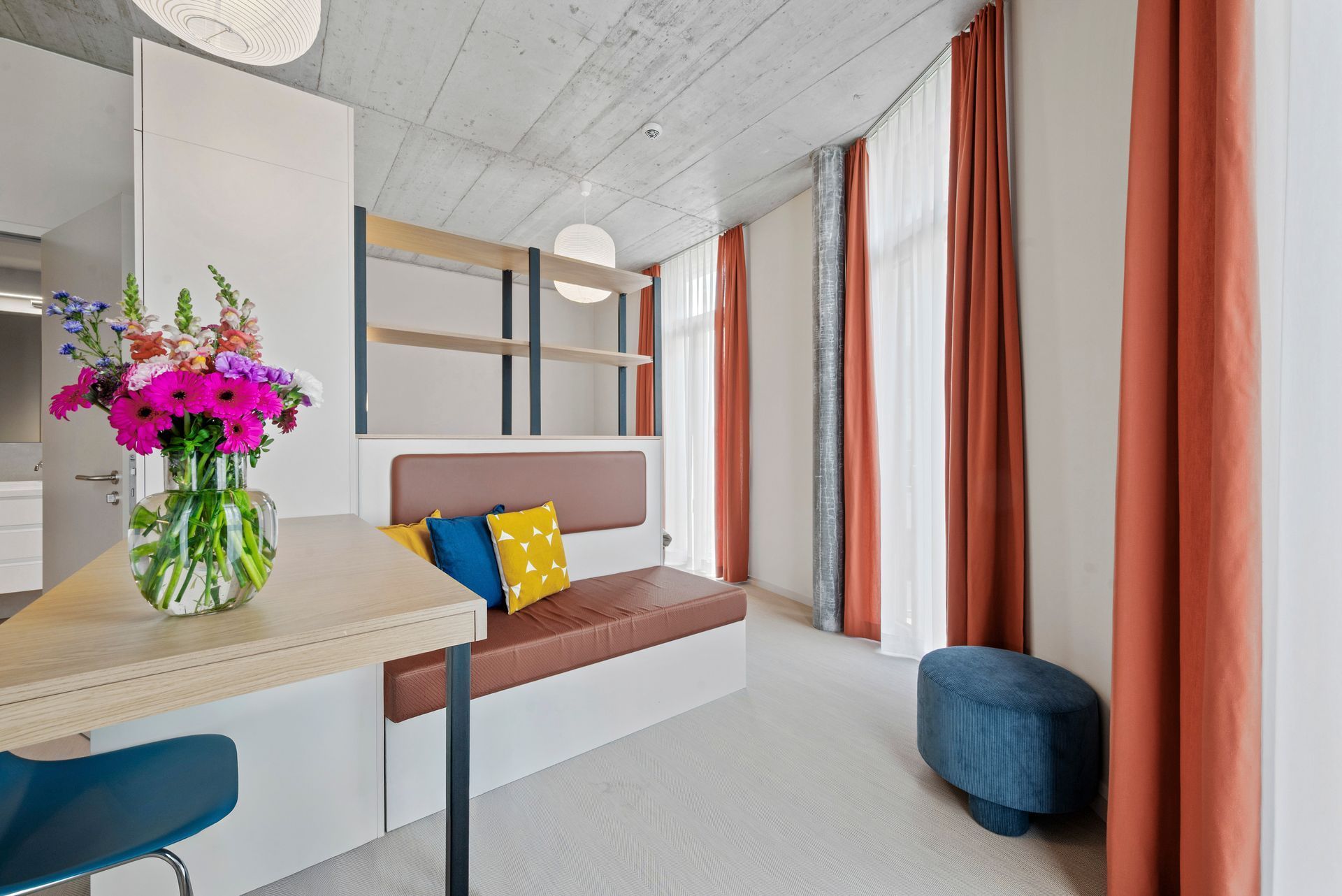 Modern living room with orange curtains, white sofa, blue ottoman, and a vase of pink flowers on a table