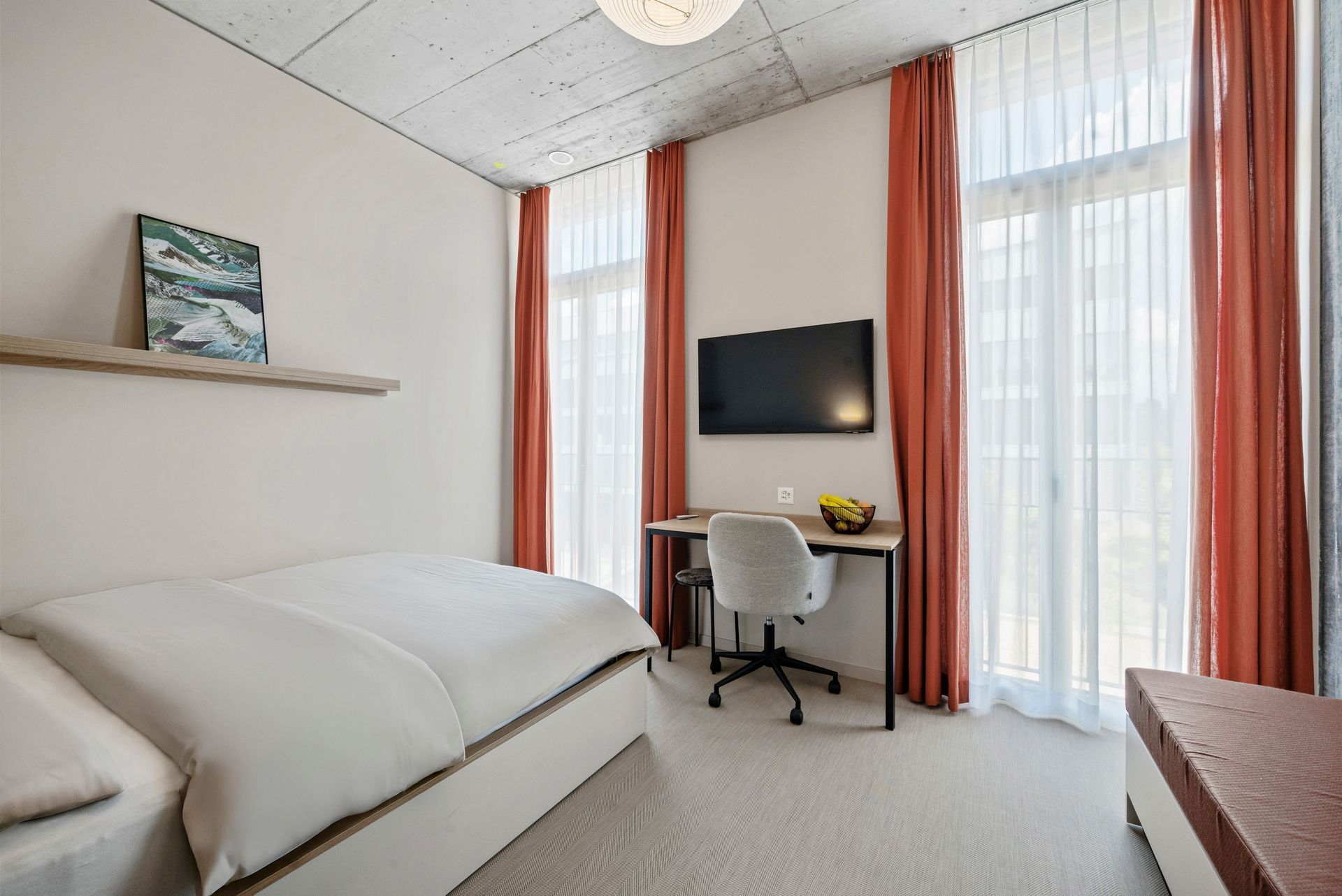 Bright bedroom with twin beds, desk, wall-mounted TV, and coral curtains by tall windows.