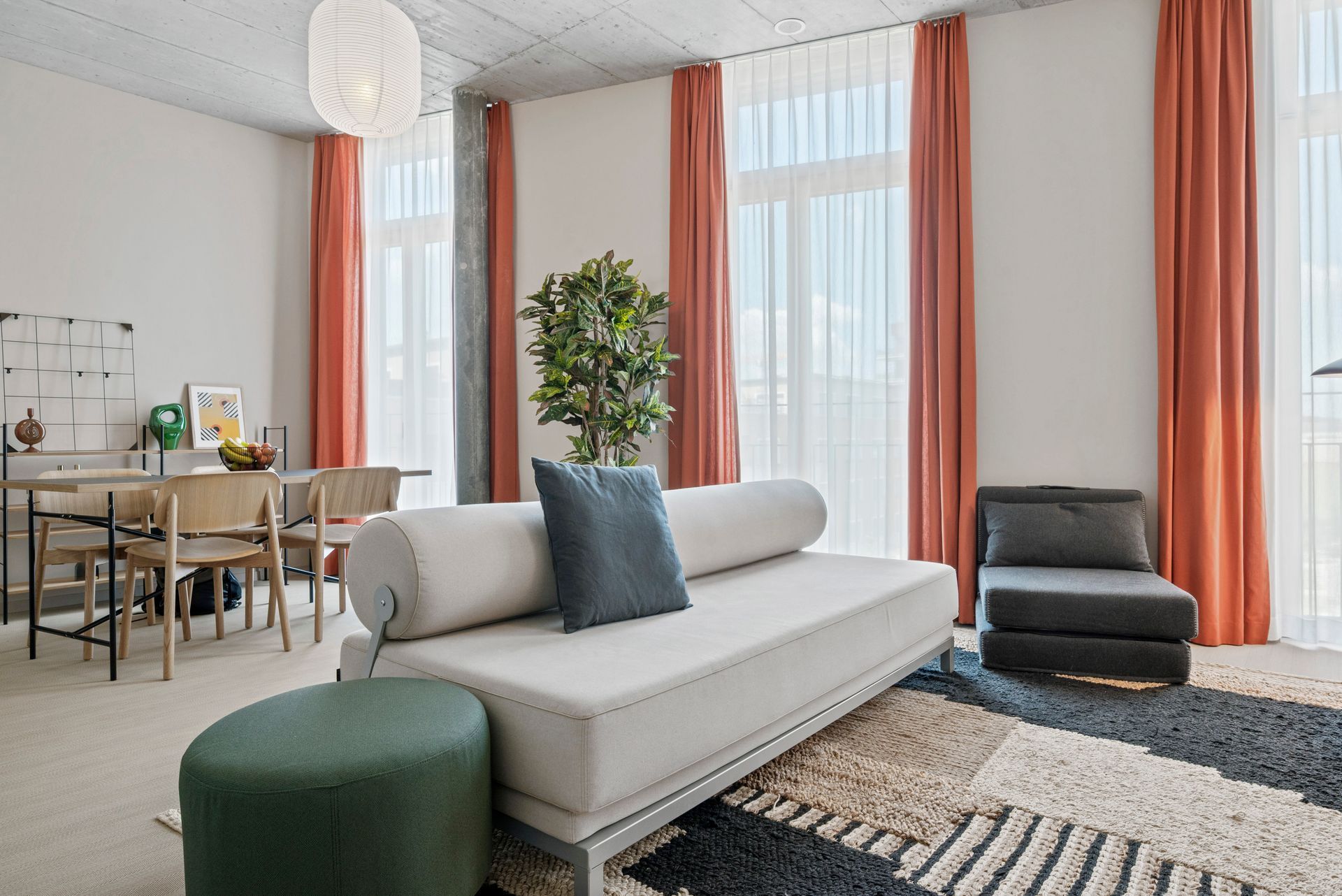 Bright living room with light sofa, dark armchair, coral curtains, rug, and dining area in the background