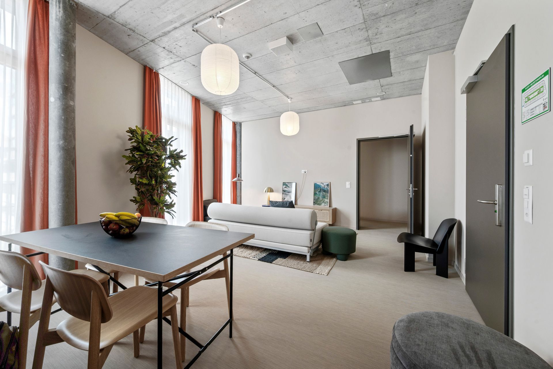 Modern living room with sofa, dining table, tall windows, plants, and exposed concrete ceiling