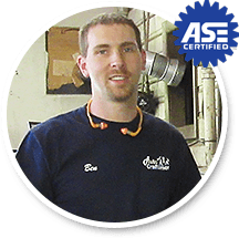 A man wearing a dark blue t-shirt and earplugs around his neck, with an ASE Certified emblem in the top right corner.