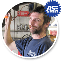 A mechanic in a blue shirt works in an auto shop with an ASE Certified emblem in the corner.