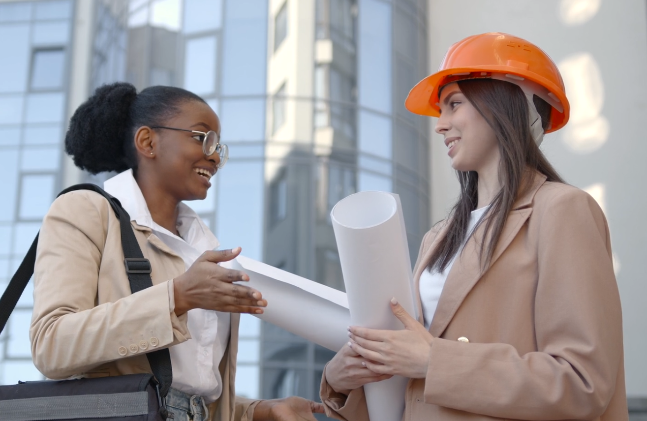 Two women in hard hats are talking to each other while holding blueprints.