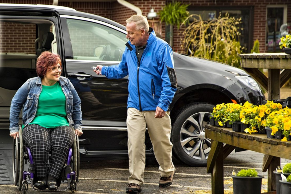 Woman in wheelchair smiles at man opening accessible van door at a plant nursery.