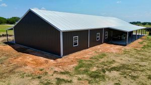 Dark brown metal-clad building with a white metal roof, porch, and windows. Set outdoors on a dry, grassy plot under a blue sky.