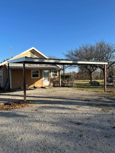 A single-story, light brown building with a carport in a gravel lot under a blue sky.
