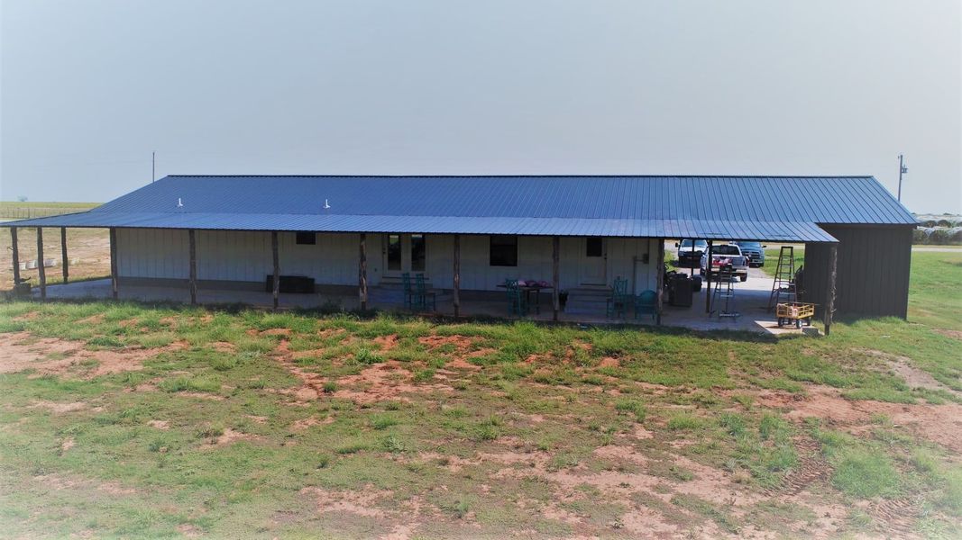 Long, single-story building with a blue metal roof and covered porch in a grassy field.