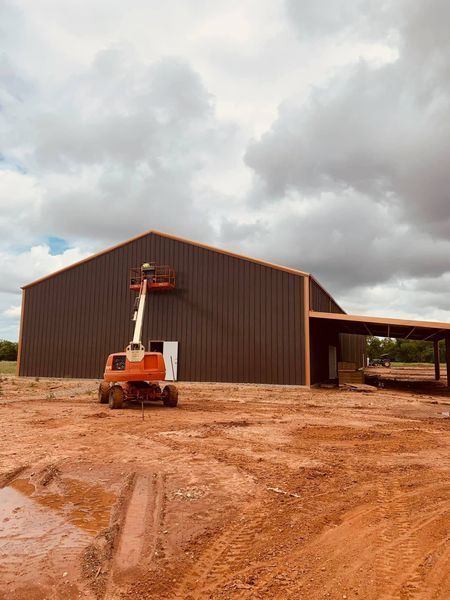 Brown metal barn under construction with lift and worker. Cloudy sky.
