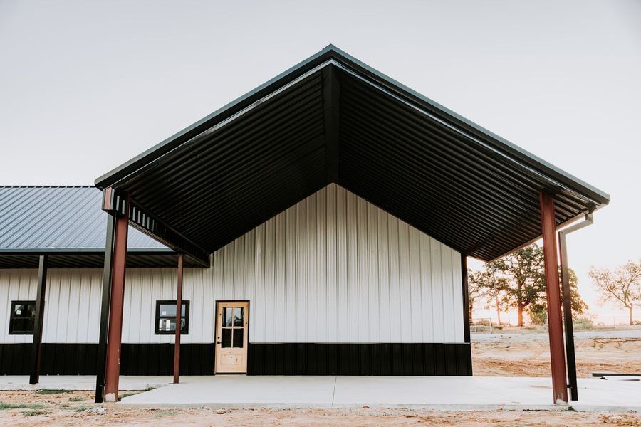White metal-sided building with black trim, under a dark corrugated metal awning supported by red poles, in an outdoor setting.