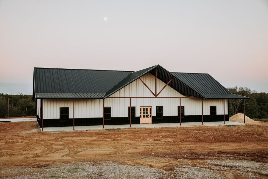 Barn-style building with black roof and trim, white siding, and a partially covered porch on a rural landscape at dusk.