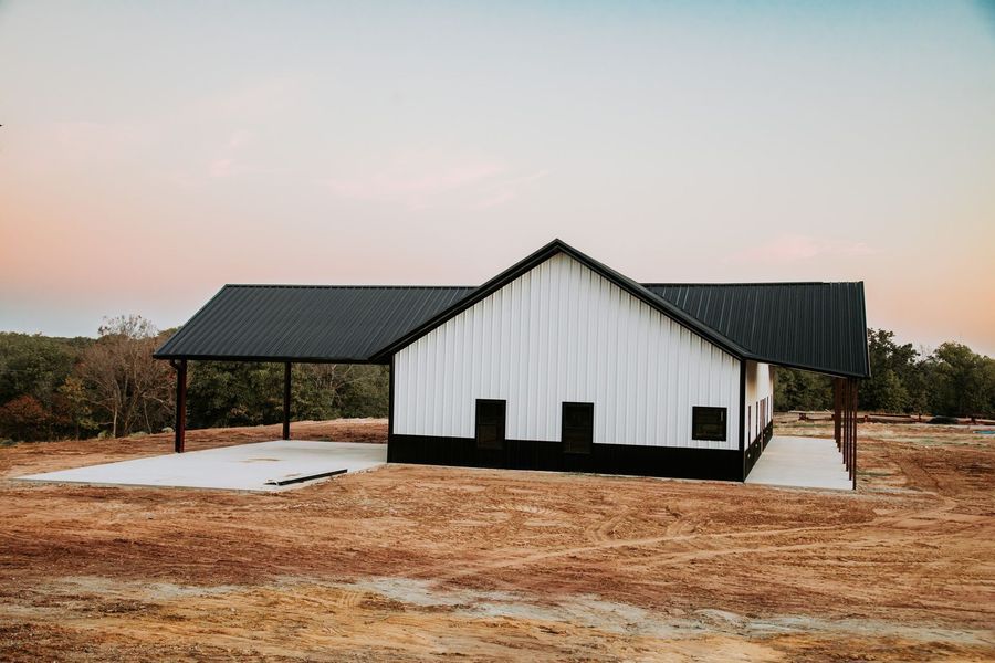 White and black barn with carport on a concrete slab in a field under a dusk sky.