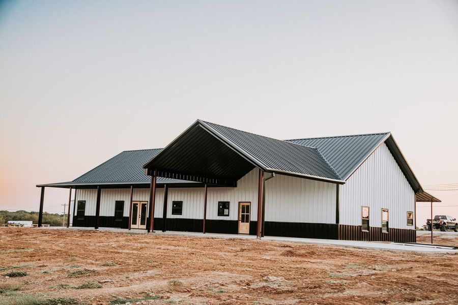 White and black barn-style building with a dark metal roof, set on a rural property under a pale sky.