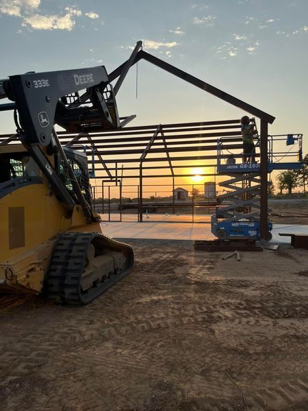 Construction of a steel-framed building at sunset; John Deere skid steer and workers on a lift.