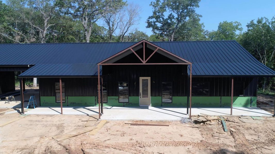 Dark blue metal-roofed building with brown trim and exposed porch on a concrete slab in a wooded area.